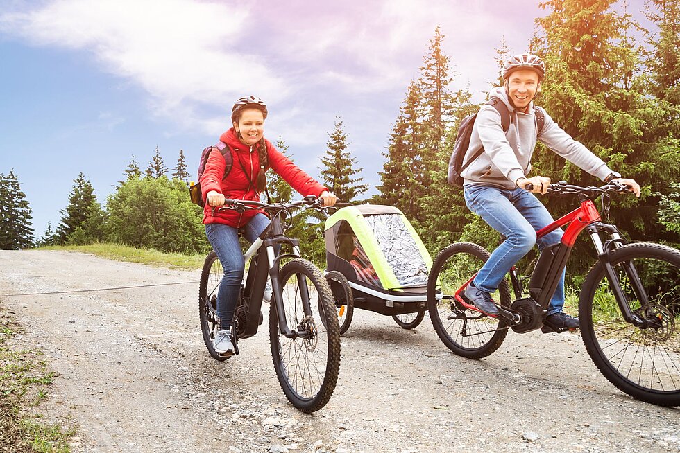 Eine Familie fährt mit E-Bikes und Kinderanhänger auf einem Schotterweg durch die Natur.